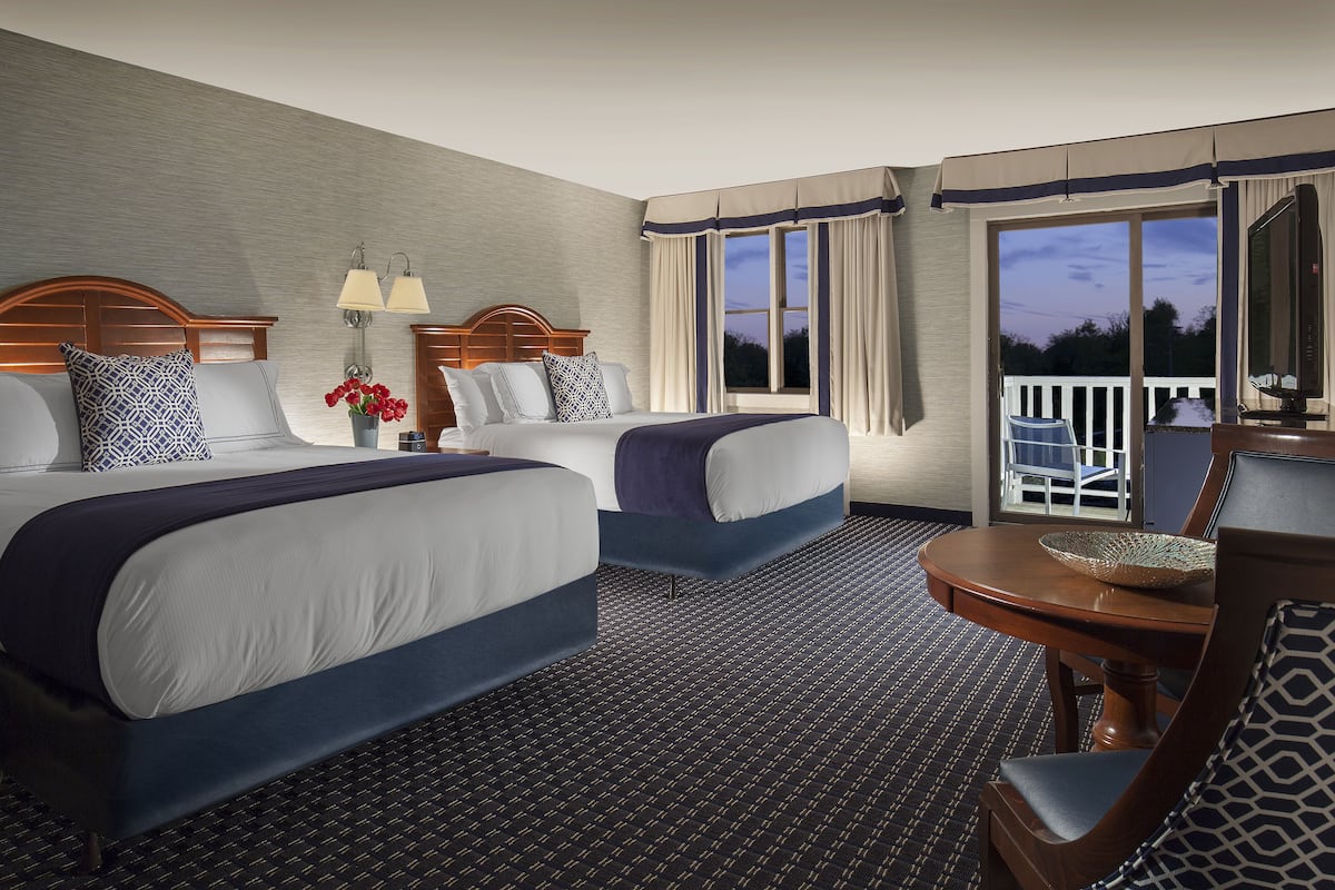 A hotel room with two beds, a small table, a chair, a TV, and a balcony with a railing.