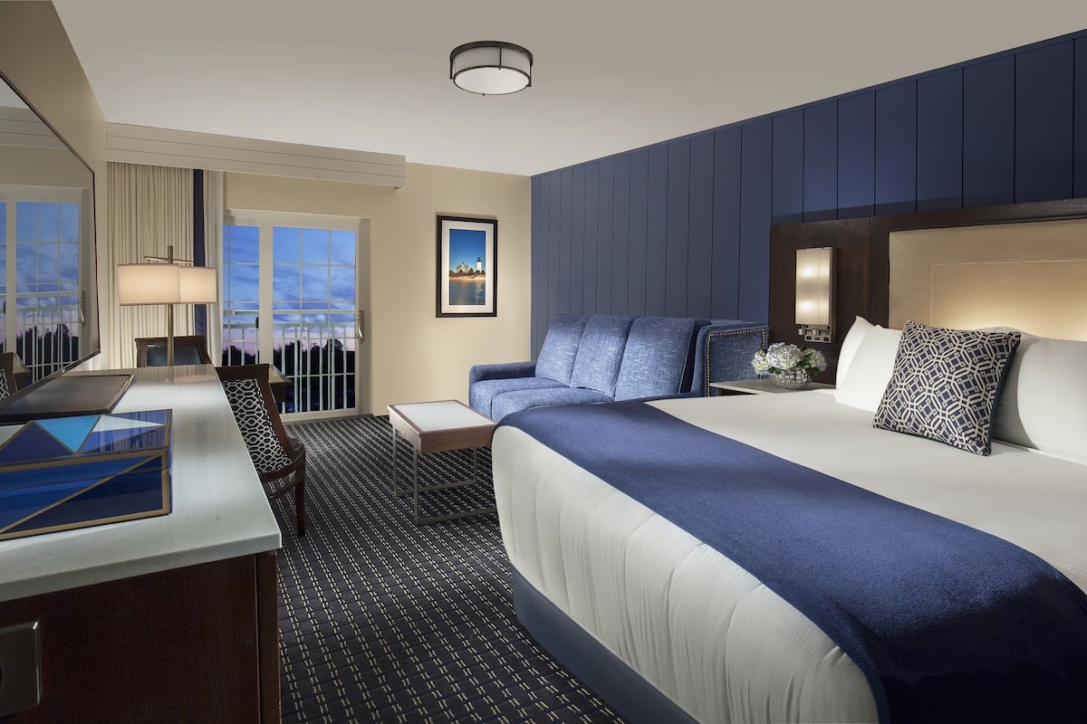 A hotel room with a large bed, a desk, a chair, a lamp, and a view of the cityscape.