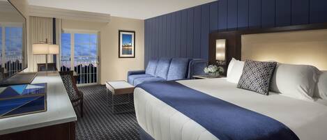 A hotel room with a large bed, a desk, a chair, a lamp, and a view of the cityscape.