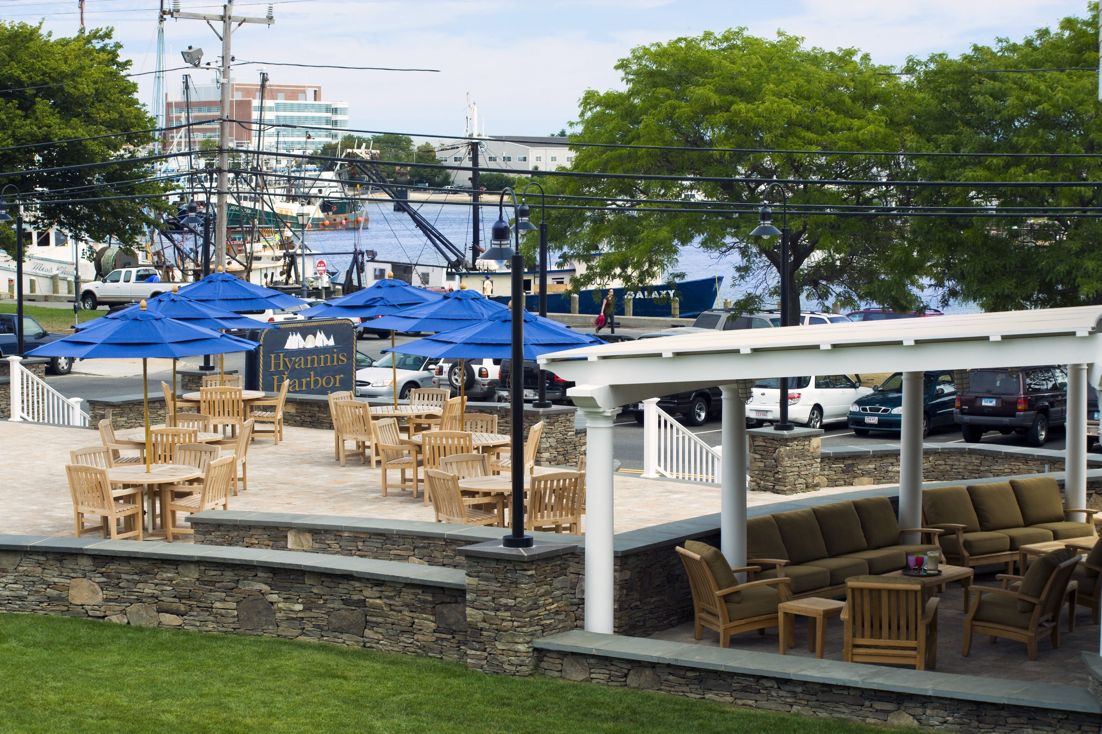 An outdoor seating area with wooden tables and chairs, covered by blue umbrellas, and a white pergola structure.