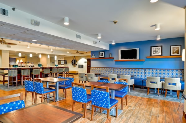 A hotel dining area with multiple tables and chairs, a bar, and a seating area with a patterned wall.