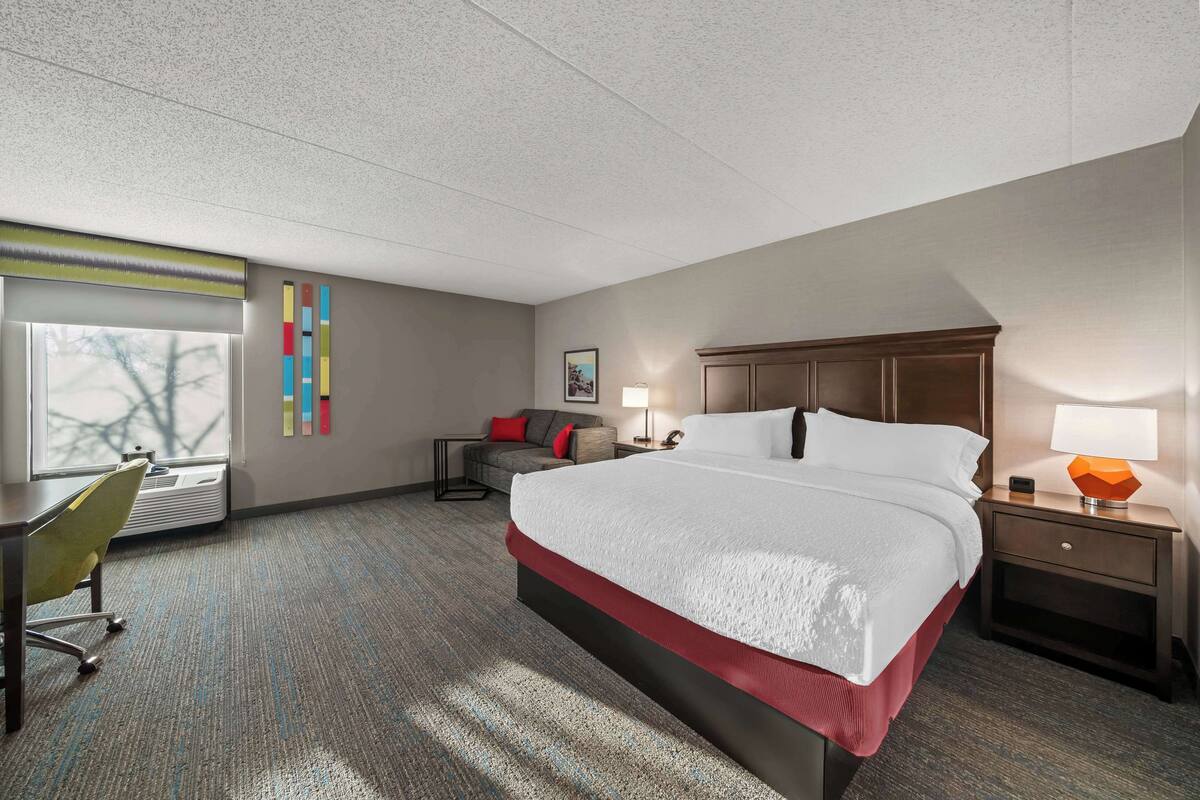 A hotel room with a large bed, a desk, a chair, a nightstand, and a window with blinds.