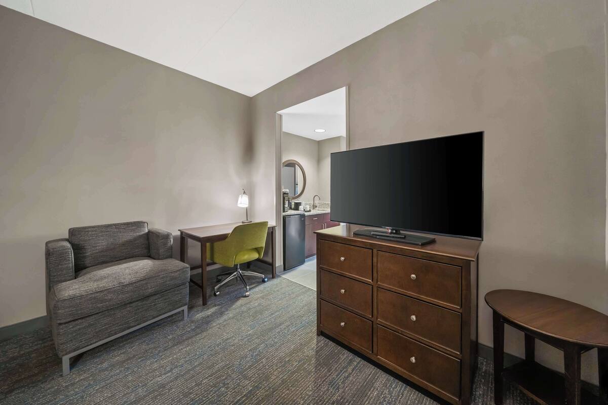A hotel room with a sofa, a desk with a chair, a dresser, a television, and a mirror.