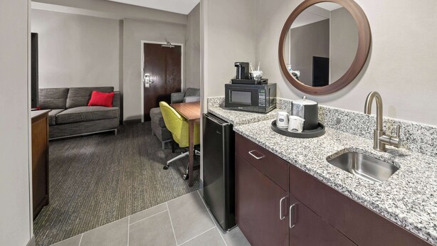 A compact hotel room with a kitchenette, a dining area with a yellow chair, a gray sofa with a red pillow, and a large mirror above the sink. - Hampton Inn by Hilton New Bedford/Fairhaven (Fairhaven)