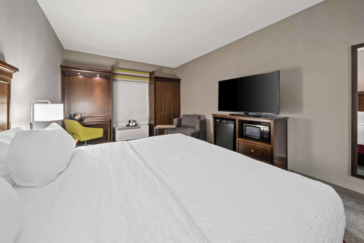 A hotel room with a large bed, a flat-screen TV, a desk with a lamp, a chair, and a wardrobe.