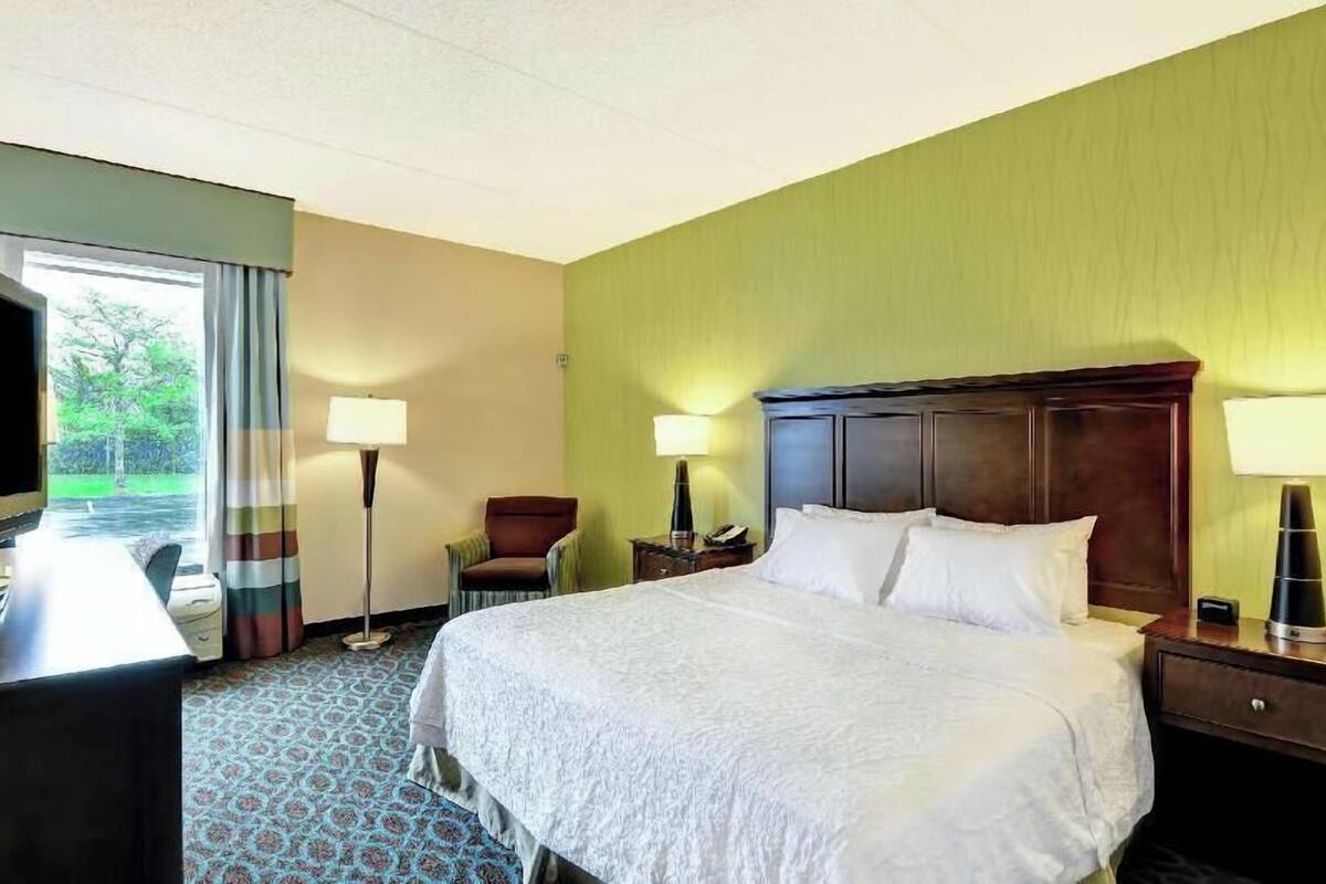 A hotel room with a bed, two bedside tables, a chair, a desk, a TV, and a window with curtains.