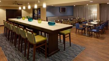 A hotel dining area with a long communal table, multiple smaller tables, and chairs.