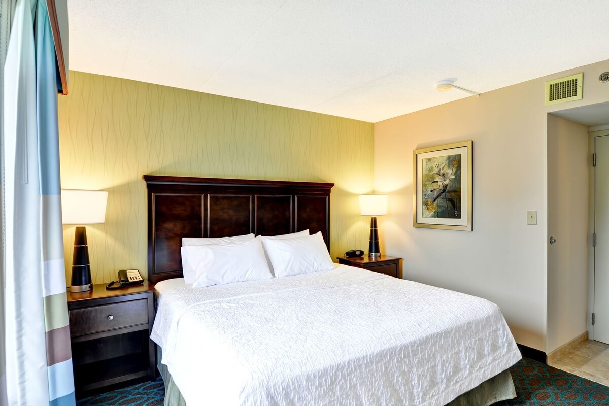 A hotel room with a large bed, two bedside tables with lamps, a painting on the wall, and a door to the right.