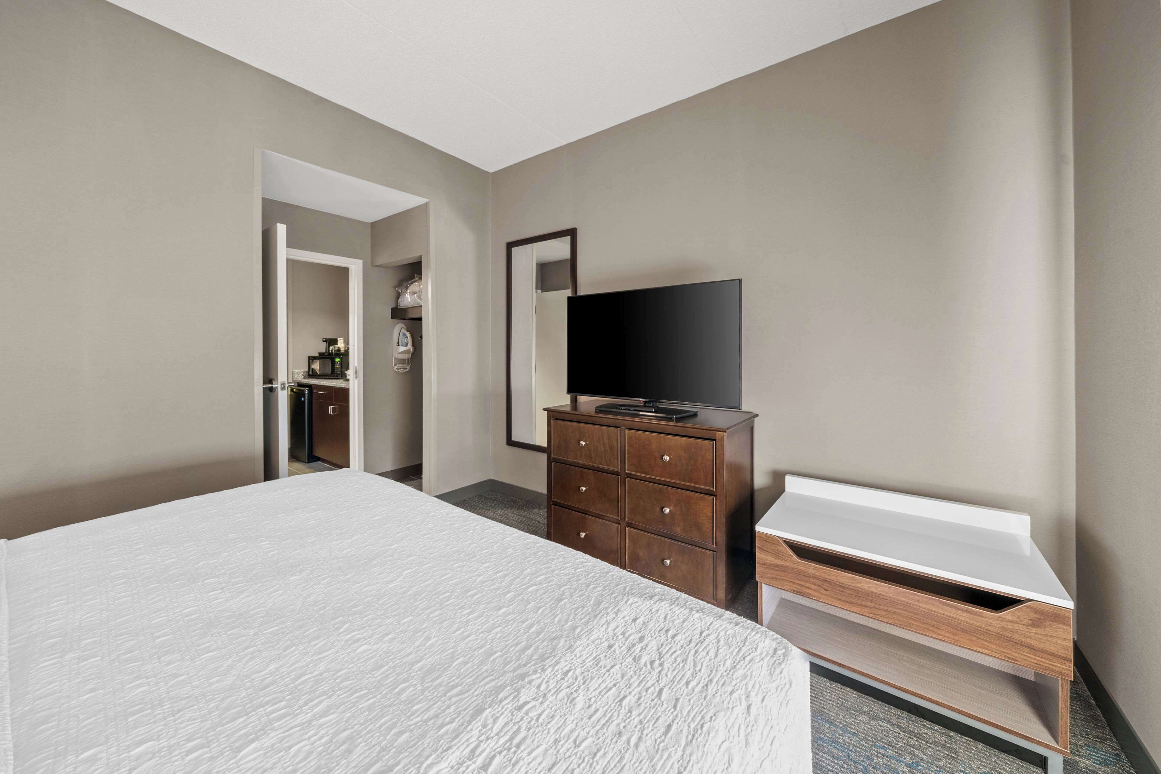 A hotel room with a large bed, a wooden dresser with a flat-screen TV, and a small desk with a white surface.