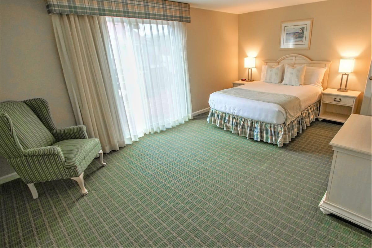 Une chambre d’hôtel comprenant un lit, une chaise, deux tables de chevet avec des lampes, une fenêtre avec des rideaux et un sol recouvert d’une moquette.