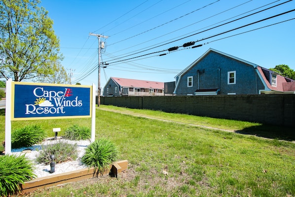A sign for Cape & Winds Resort with a grassy area and a brick wall in the background.