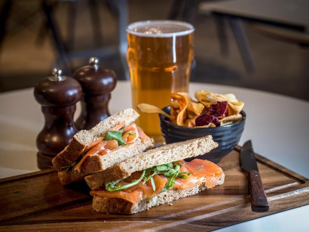 Un sándwich de salmón ahumado, una jarra de cerveza y un bol de papas fritas sobre una tabla de madera.