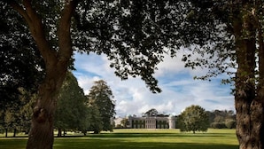A large building with a symmetrical facade and a central entrance, surrounded by a well-maintained lawn and mature trees. - The Goodwood Hotel (Chichester)