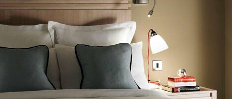 A neatly made bed with white and dark gray pillows, a wooden headboard, a bedside lamp, and a small bedside table with books and a decorative sphere.