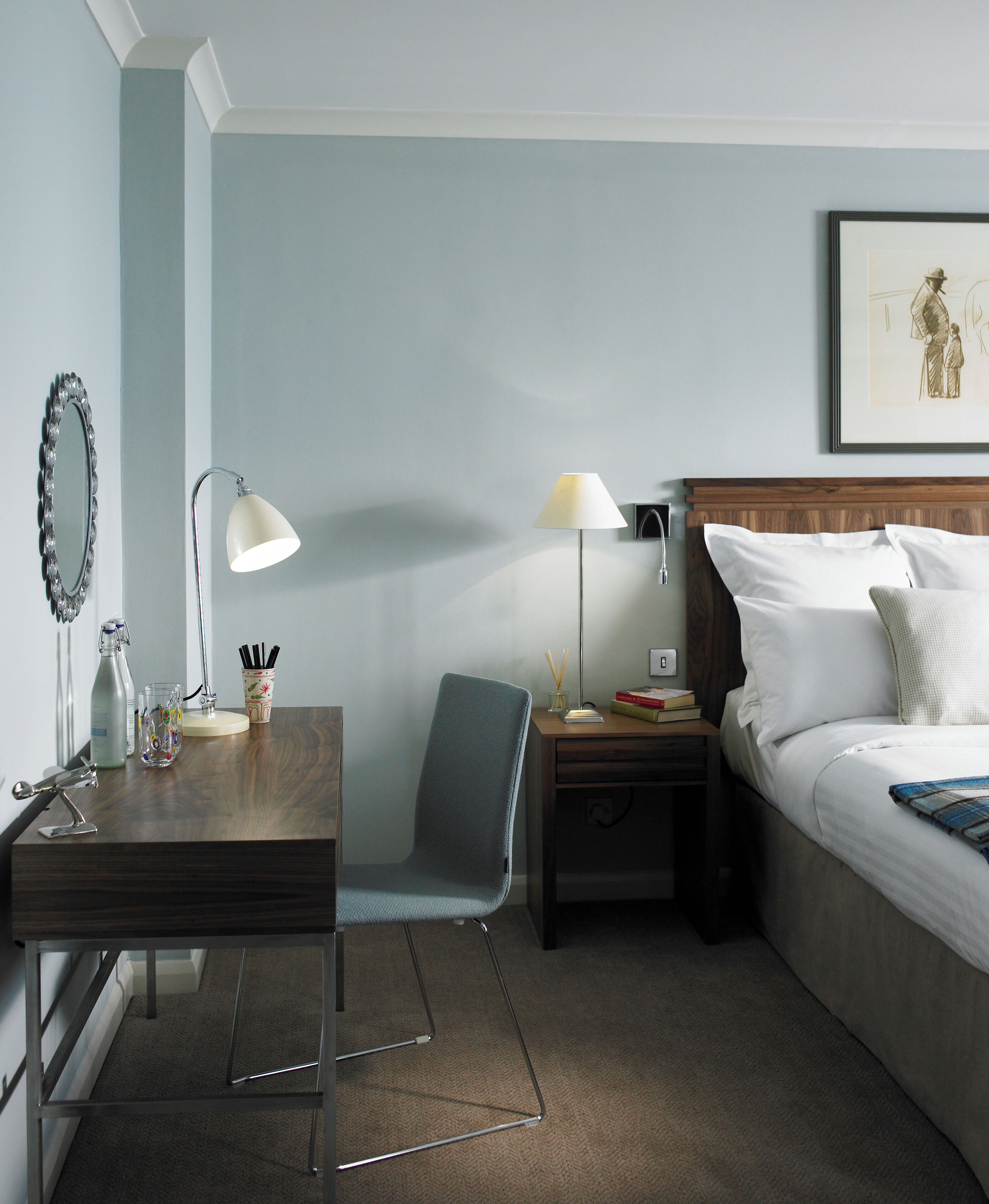 Een hotelkamer met een houten bureau, een stoel, een bed met witte lakens, een nachtkastje, een lamp, een ingelijst schilderij aan de muur en een spiegel.