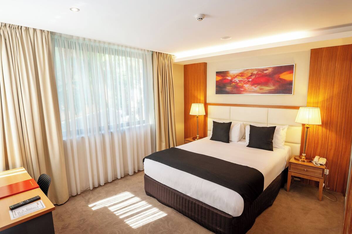 A hotel room with a large bed, two bedside lamps, a desk with a telephone, and a window with curtains.