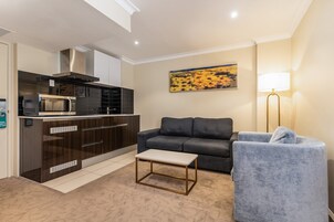 Un espacio habitable compacto con kitchenette, un sofá gris, una mesita y una lámpara de pie.
