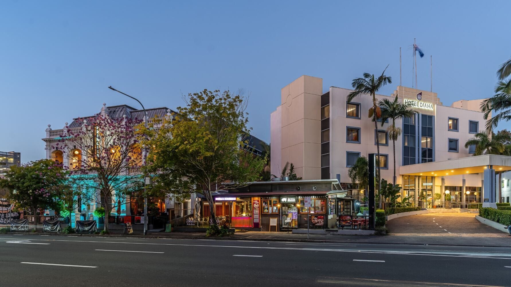 Un hotel con fachada moderna, una entrada visible y vista a la calle con un árbol y un edificio con luces de colores.