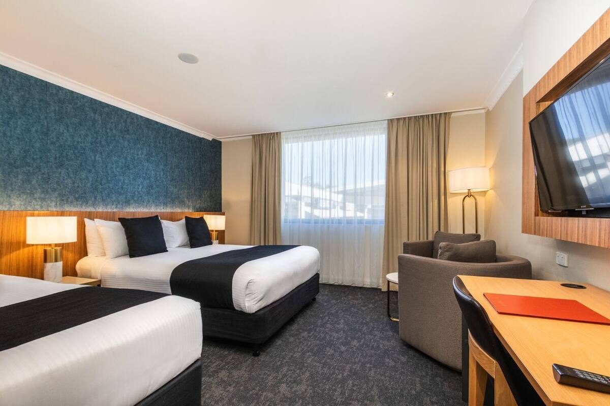 A hotel room with two beds, a desk, a chair, a TV, and a large window with curtains.