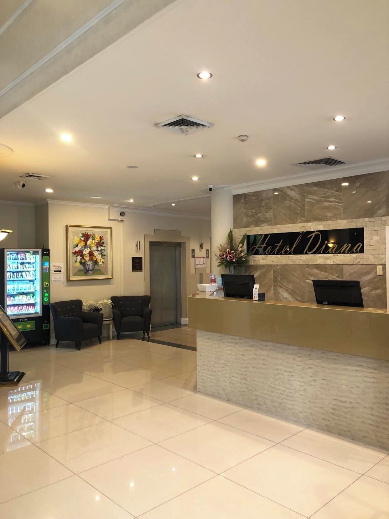 A hotel reception area with a marble counter, two black armchairs, a vending machine, and a framed floral artwork.