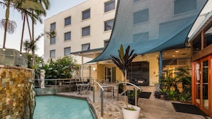 A pool area with a stone wall, a lounge with tables and chairs, and a building with multiple windows. - Hotel Diana Woolloongabba (Woolloongabba)