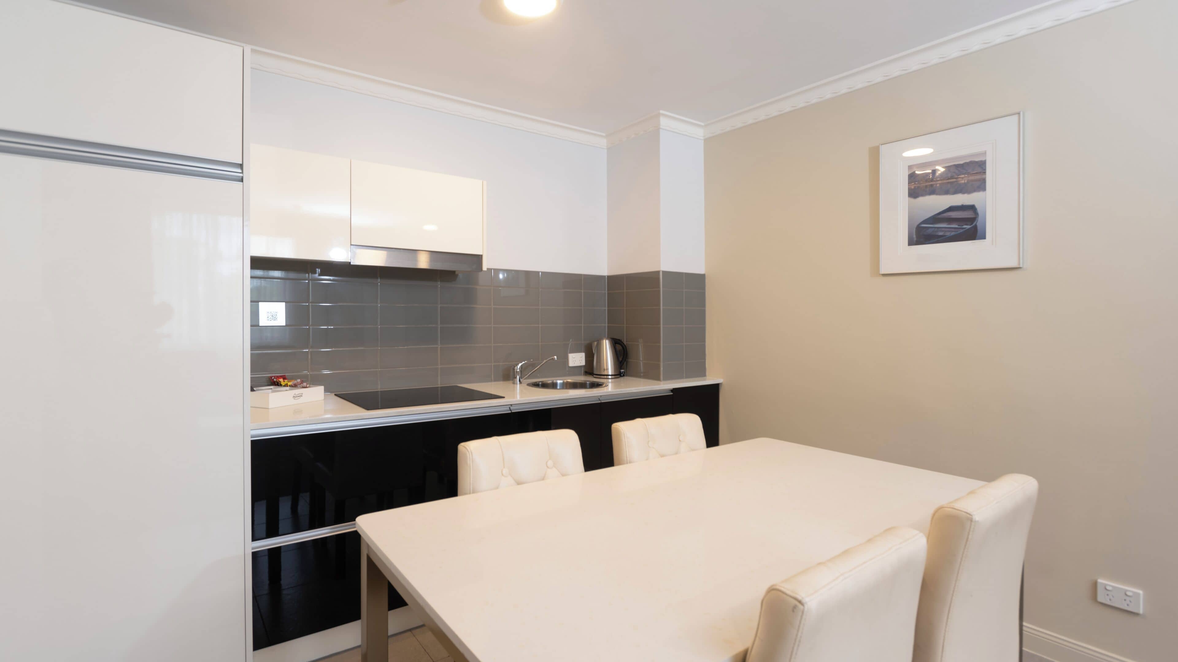 A compact kitchen with a dining area, featuring a white table and chairs, a black countertop, and a tiled backsplash.