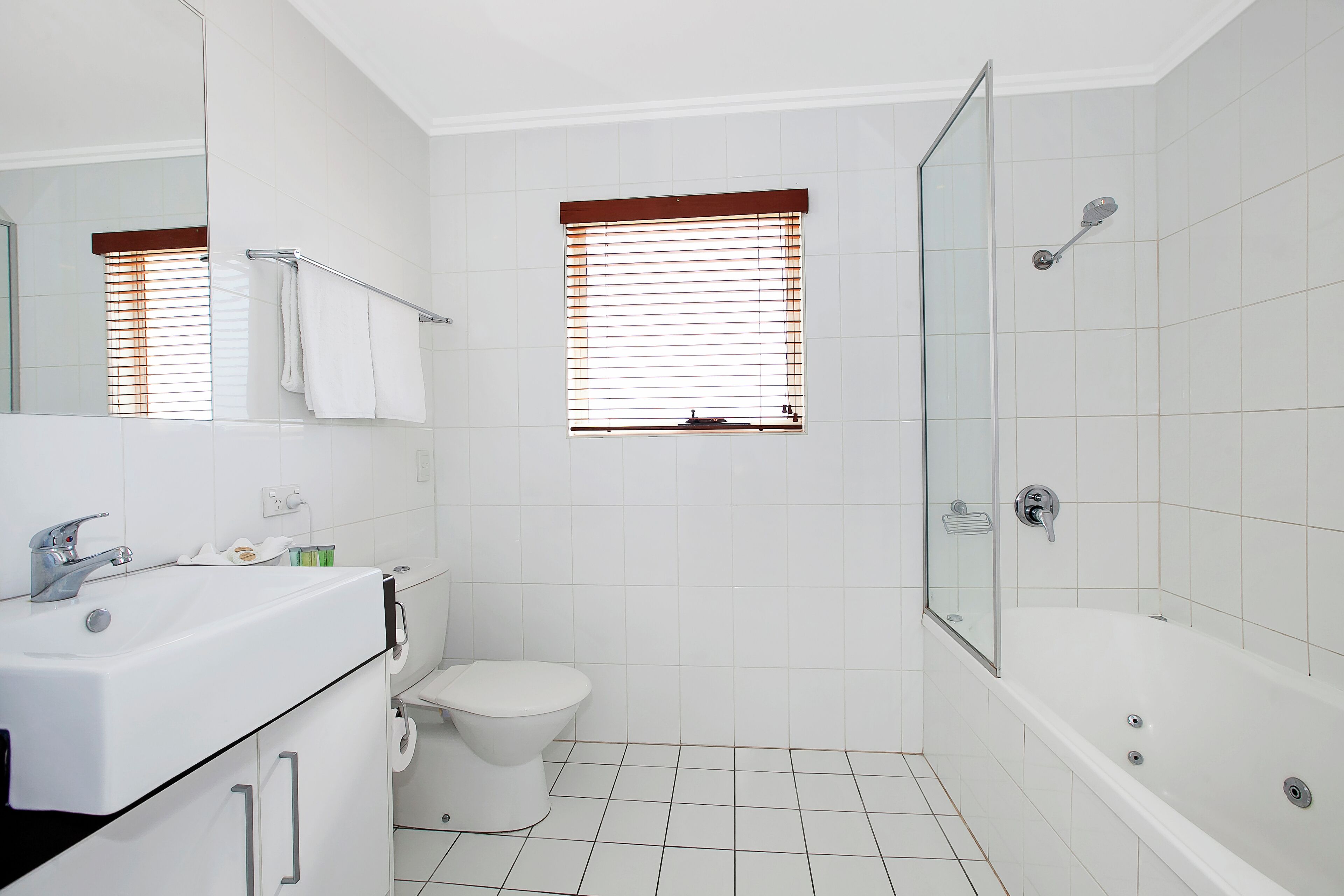 Un baño con lavamanos, inodoro y bañera blancos. Cuenta con dos ventanas con persianas y una puerta de ducha.