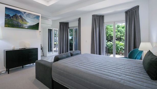 A modern bedroom with a large bed, a black dresser, a painting, and a view of greenery through the windows.