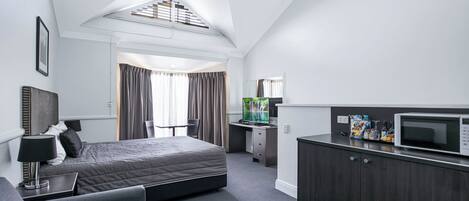 A modern hotel room with a large bed, a desk with a computer, a television, and a kitchenette.