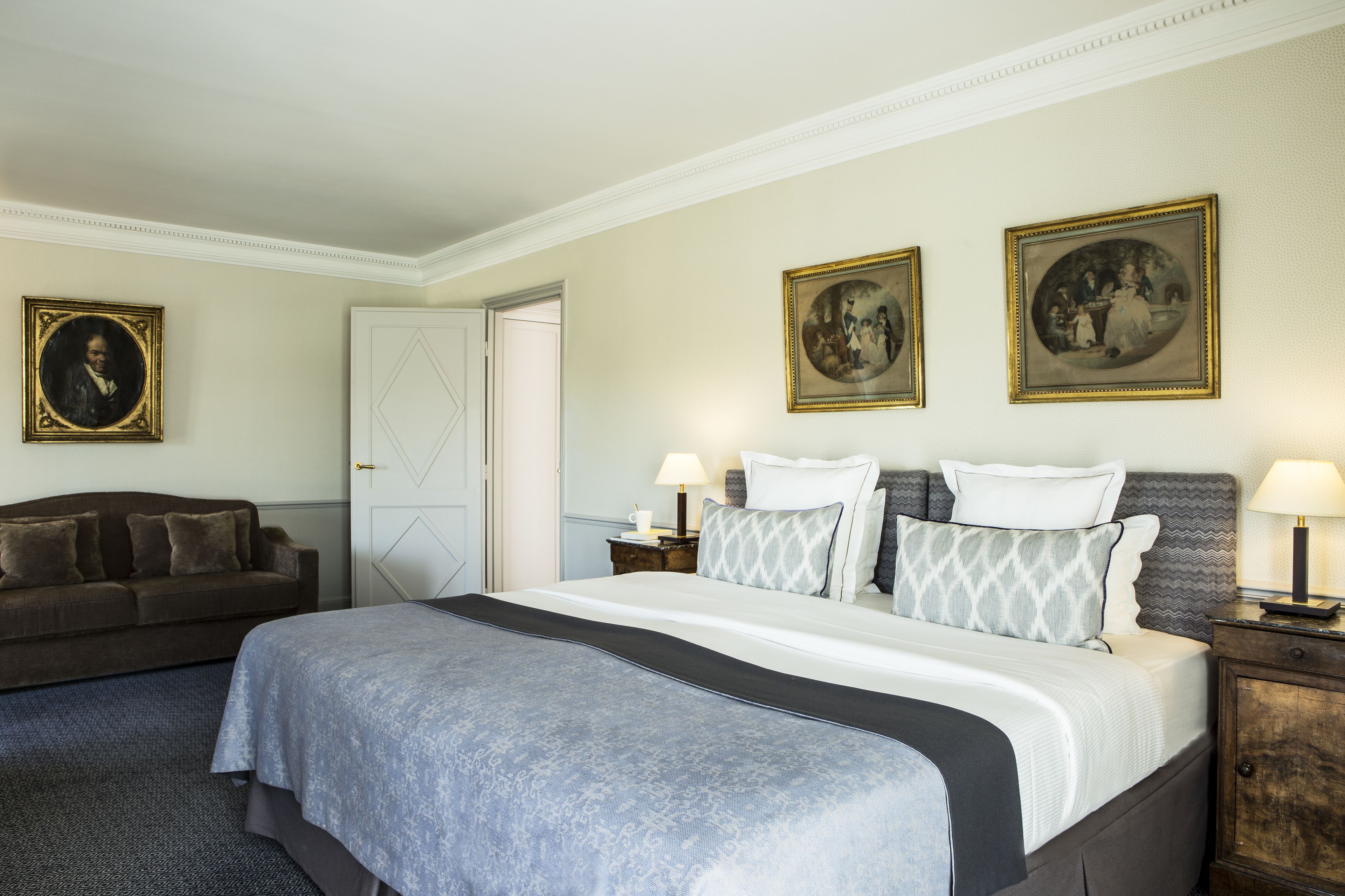 Habitación de hotel con una cama grande, un sofá, dos mesitas de noche con lámparas y dos cuadros en la pared.