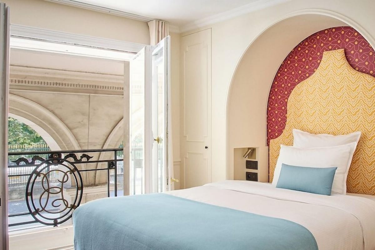 A hotel room with a bed, an arched headboard, a balcony with a railing, and a view of greenery outside.