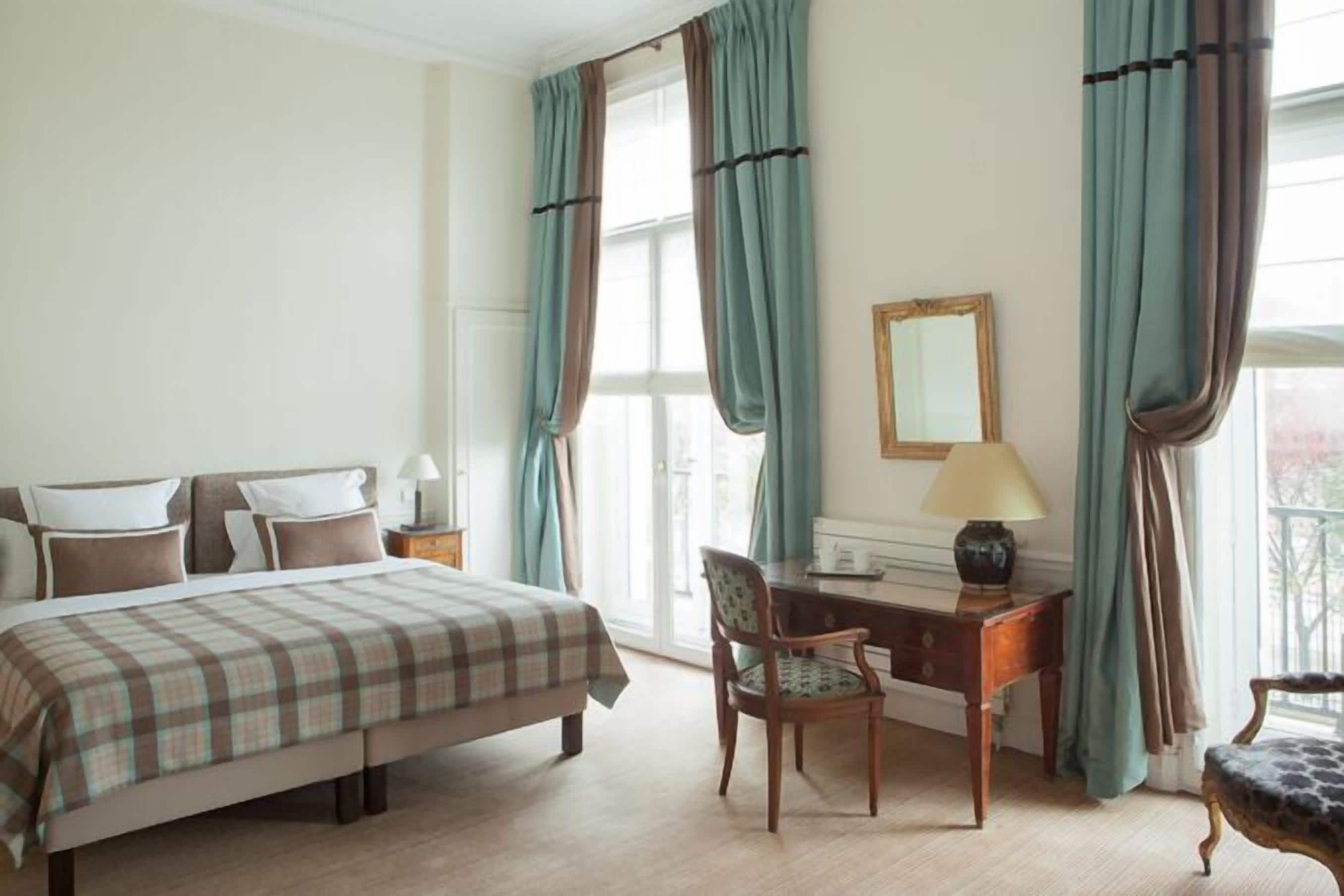 A hotel room with a bed, a desk, a chair, a lamp, and curtains.