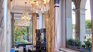 A luxurious hotel lobby with a marble floor, a large sofa with blue cushions, a round glass table, and a chandelier. - Hotel Brighton (Paris)