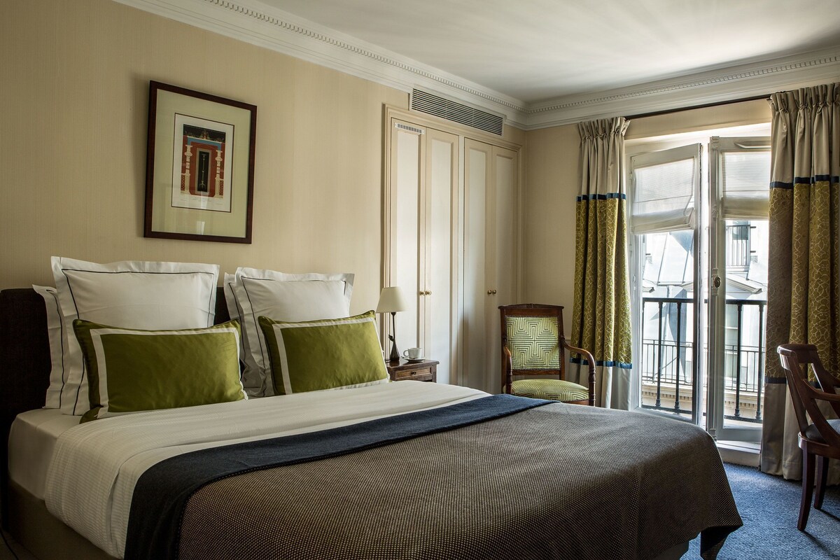 A hotel room with a large bed, a framed picture on the wall, a chair, and a balcony with curtains.