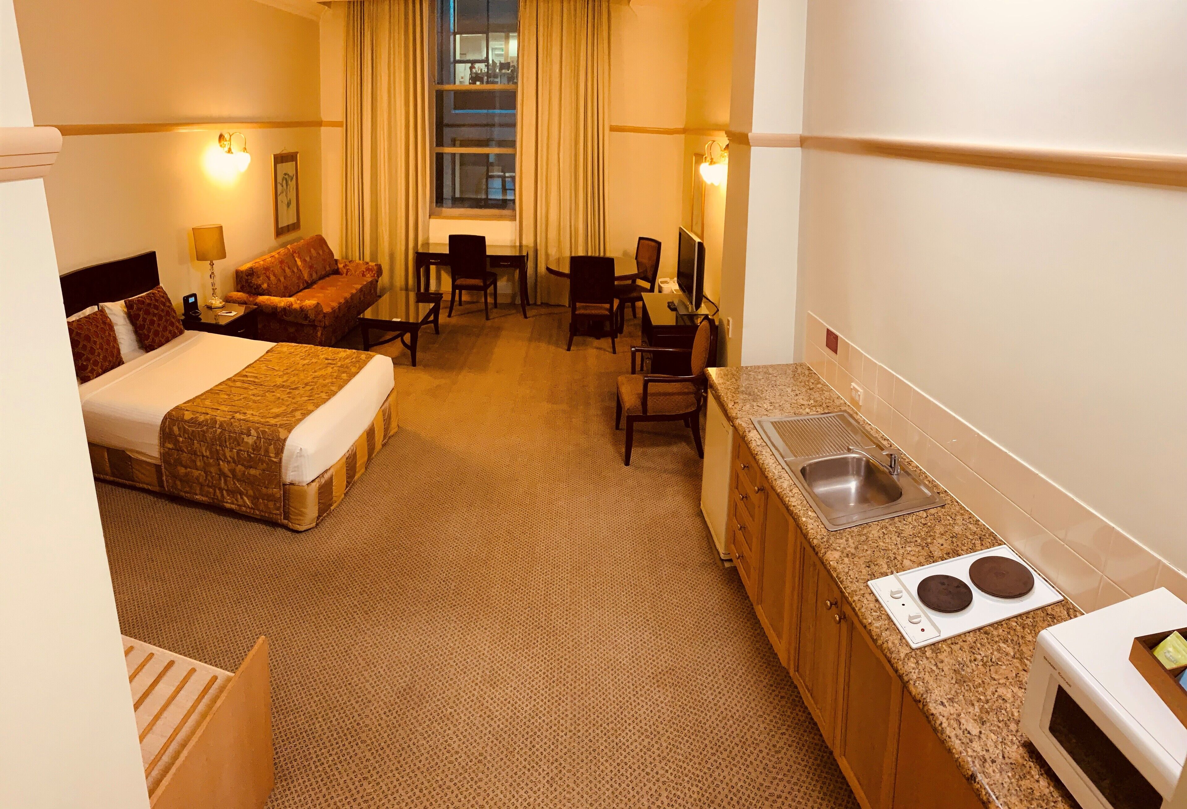 Habitación de hotel con cama, sofá, escritorio, silla, televisor y kitchenette con lavaplatos y estufa.