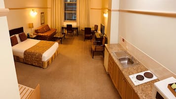 Habitación de hotel con cama, sofá, escritorio, silla, televisor y kitchenette con lavaplatos y estufa.