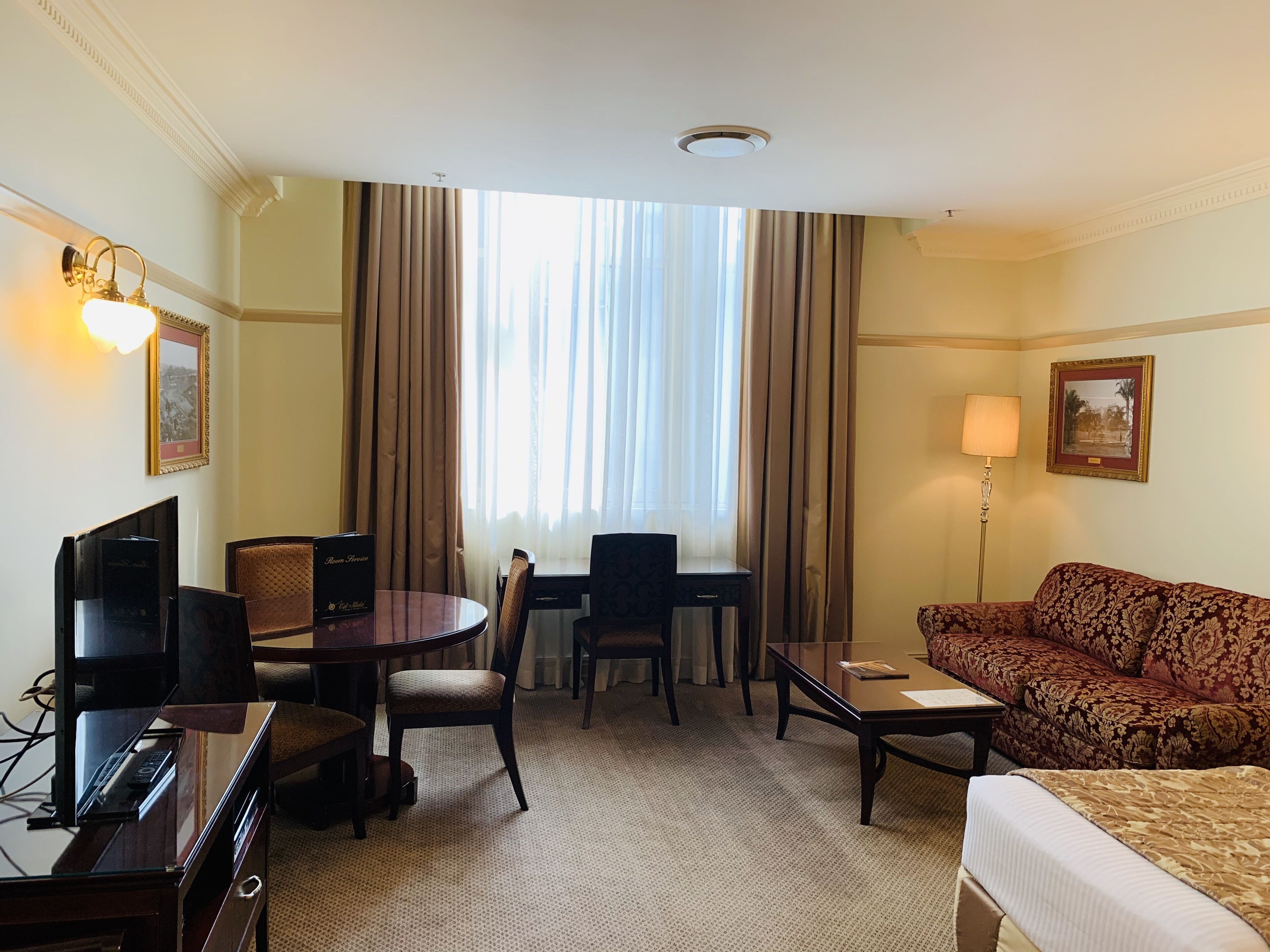 A hotel room with a bed, a desk with a computer, a TV, a sofa, and a large window with curtains.