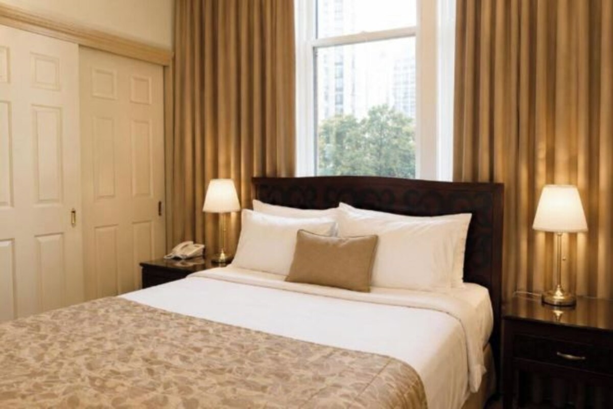 A hotel room with a large bed, two bedside tables with lamps, a window with curtains, and a door.