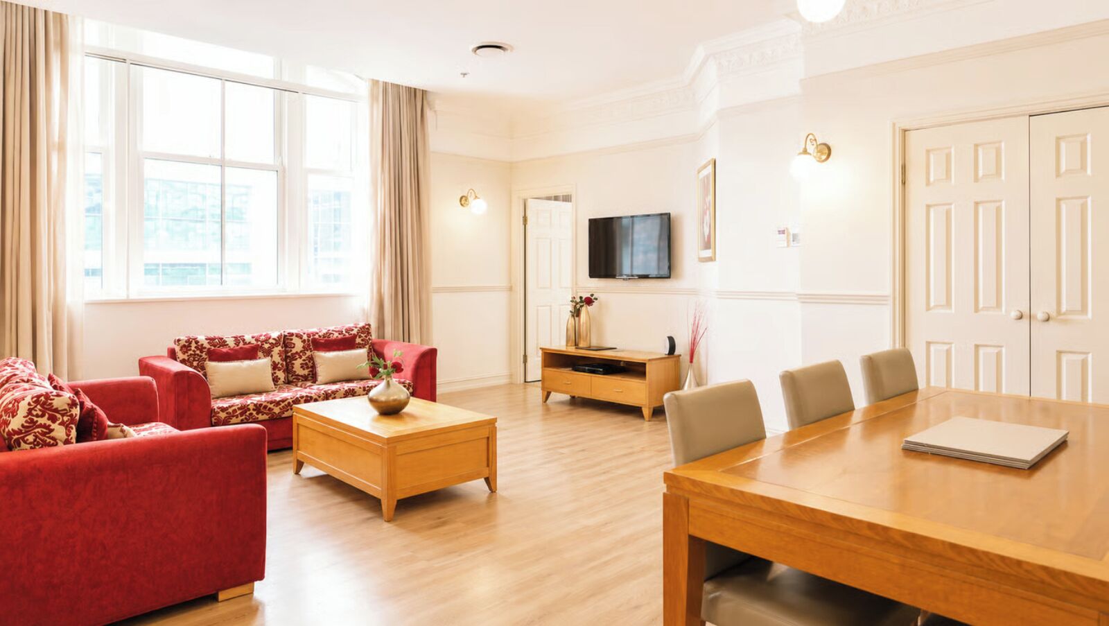 A spacious living area with a red sofa set, a wooden coffee table, a TV mounted on the wall, and a conference table with chairs.