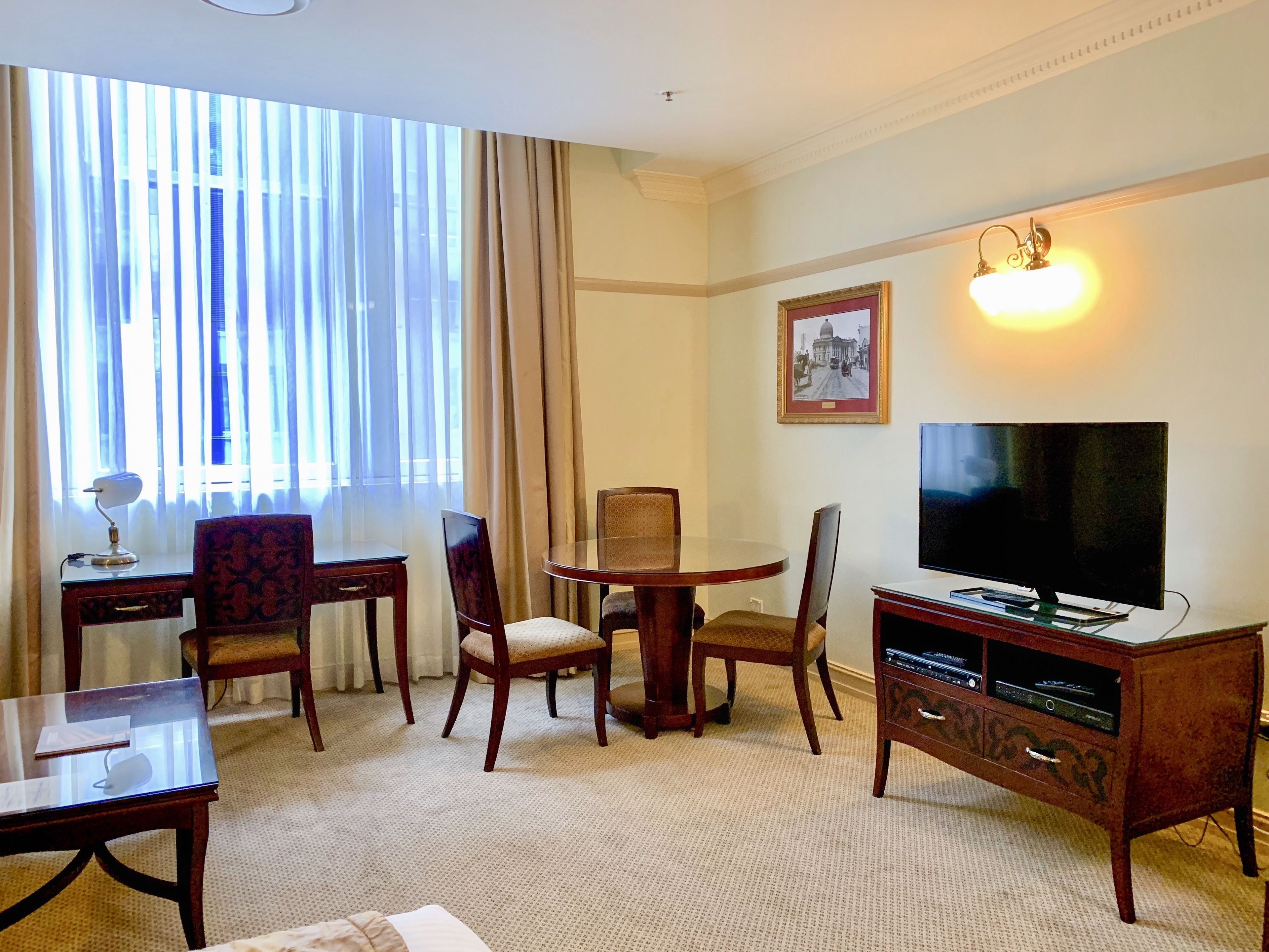 A hotel room with a large window, a television, a desk with a lamp, and a chair.