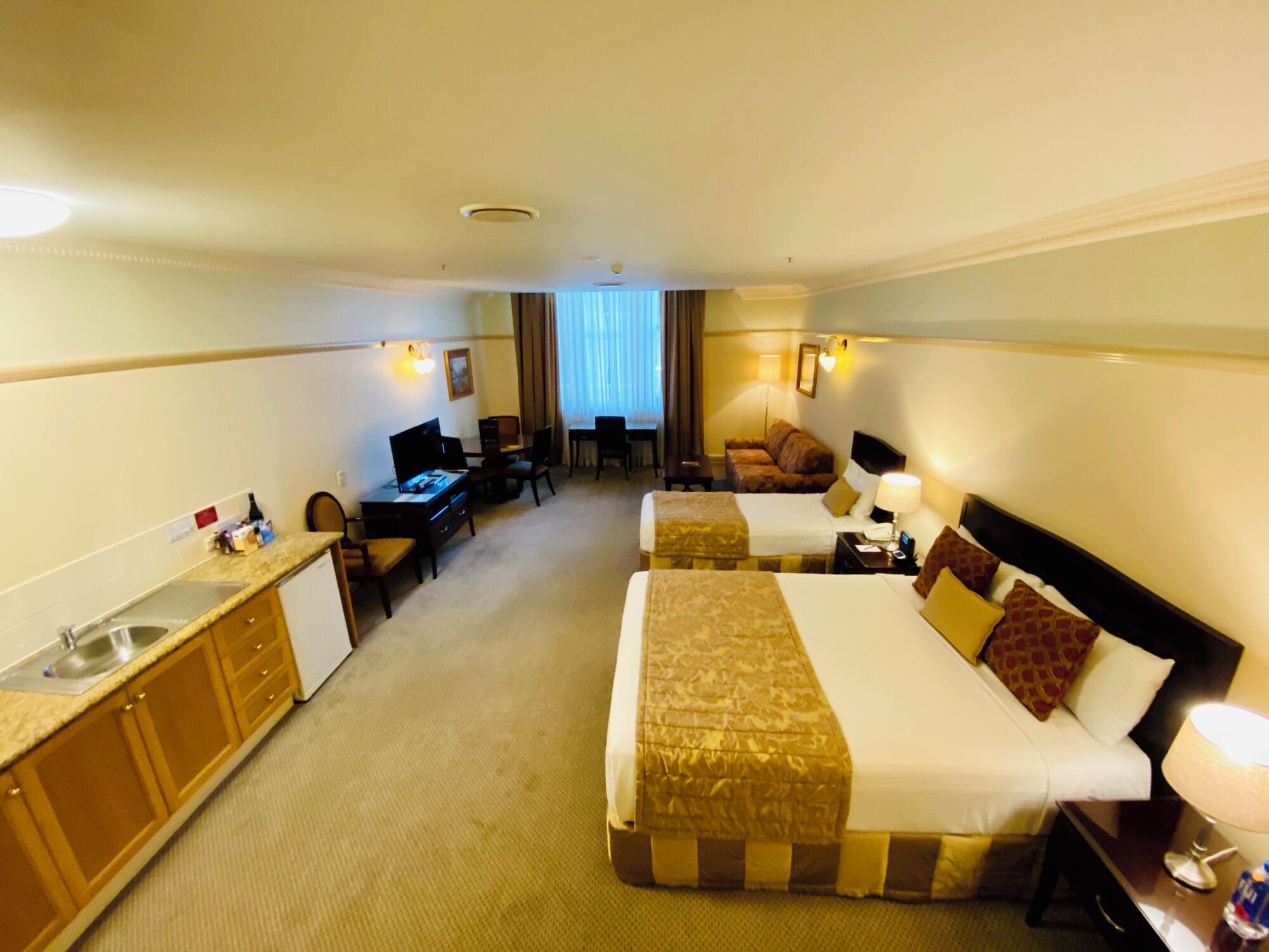 A hotel room with two beds, a desk, a chair, a small kitchenette, and a window with curtains.