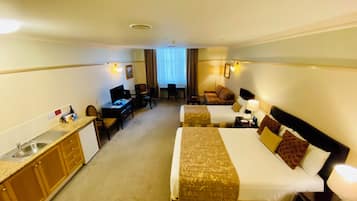A hotel room with two beds, a desk, a chair, a small kitchenette, and a window with curtains.