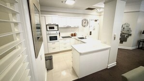 A modern kitchen with white cabinetry, a microwave, oven, and sink. There is a small trash bin and a decorative wall piece. - Royal Albert Hotel (Brisbane)
