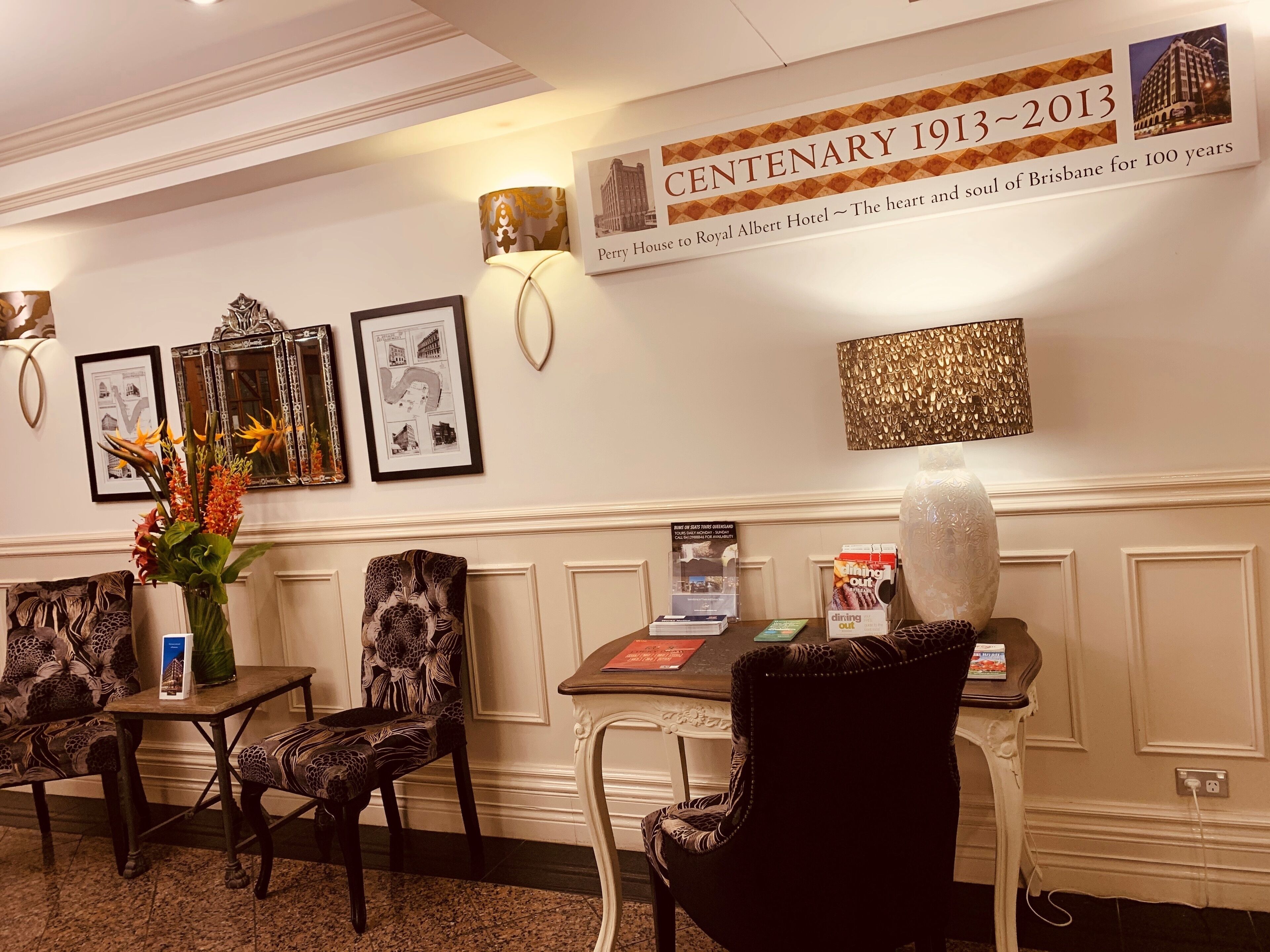 A hotel lobby with a centenary celebration banner, framed pictures, a vase with flowers, and a table with a menu.
