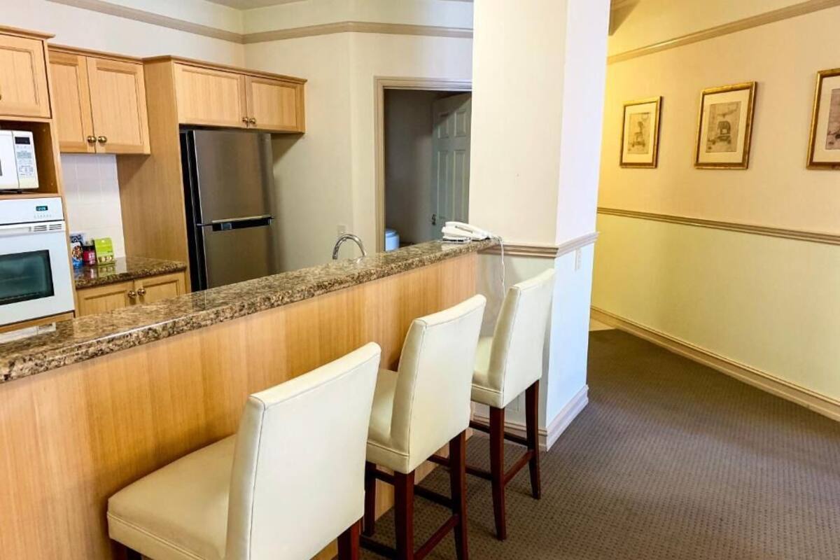 A kitchen with wooden cabinets, a granite countertop, a stainless steel refrigerator, and a bar area with white bar stools.