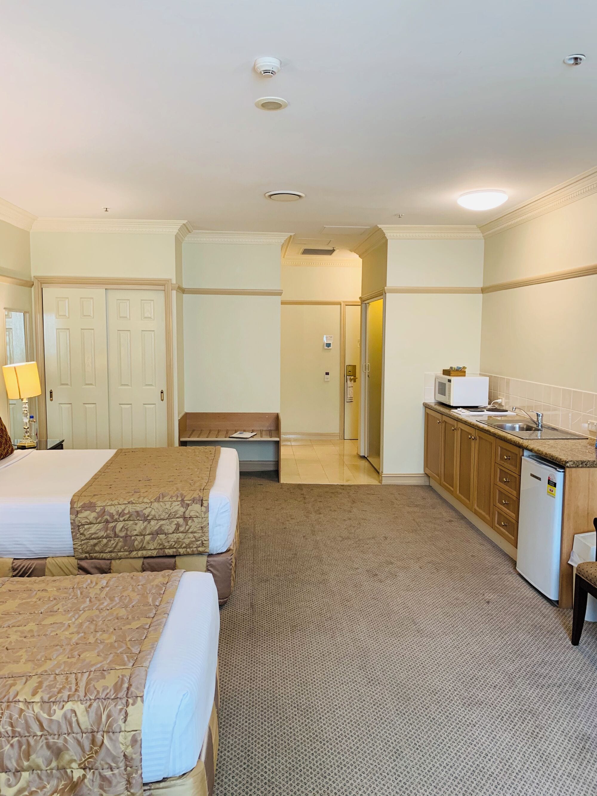 A hotel room with two beds, a small kitchenette, a bathroom, and a hallway.