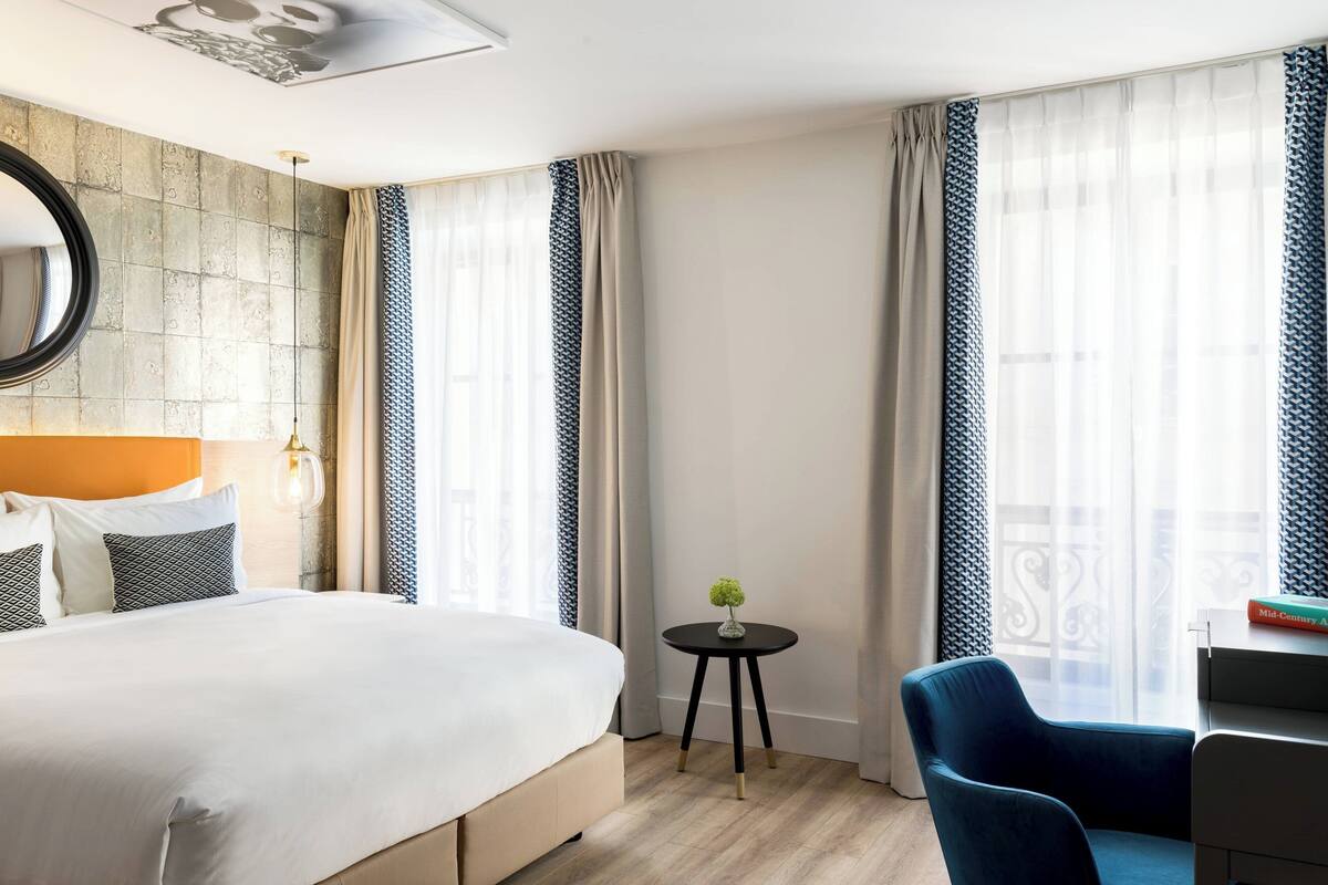 A hotel room with a large bed, a round table with a vase, a blue chair, and a view of the city through large windows.