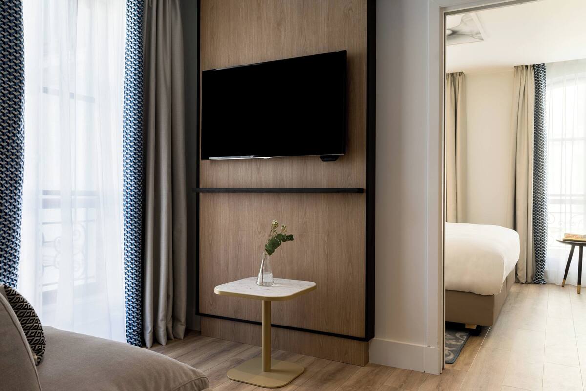 A modern hotel room with a flat-screen TV mounted on the wall, a small table with a vase, a bed, and a view of the outside through large windows.