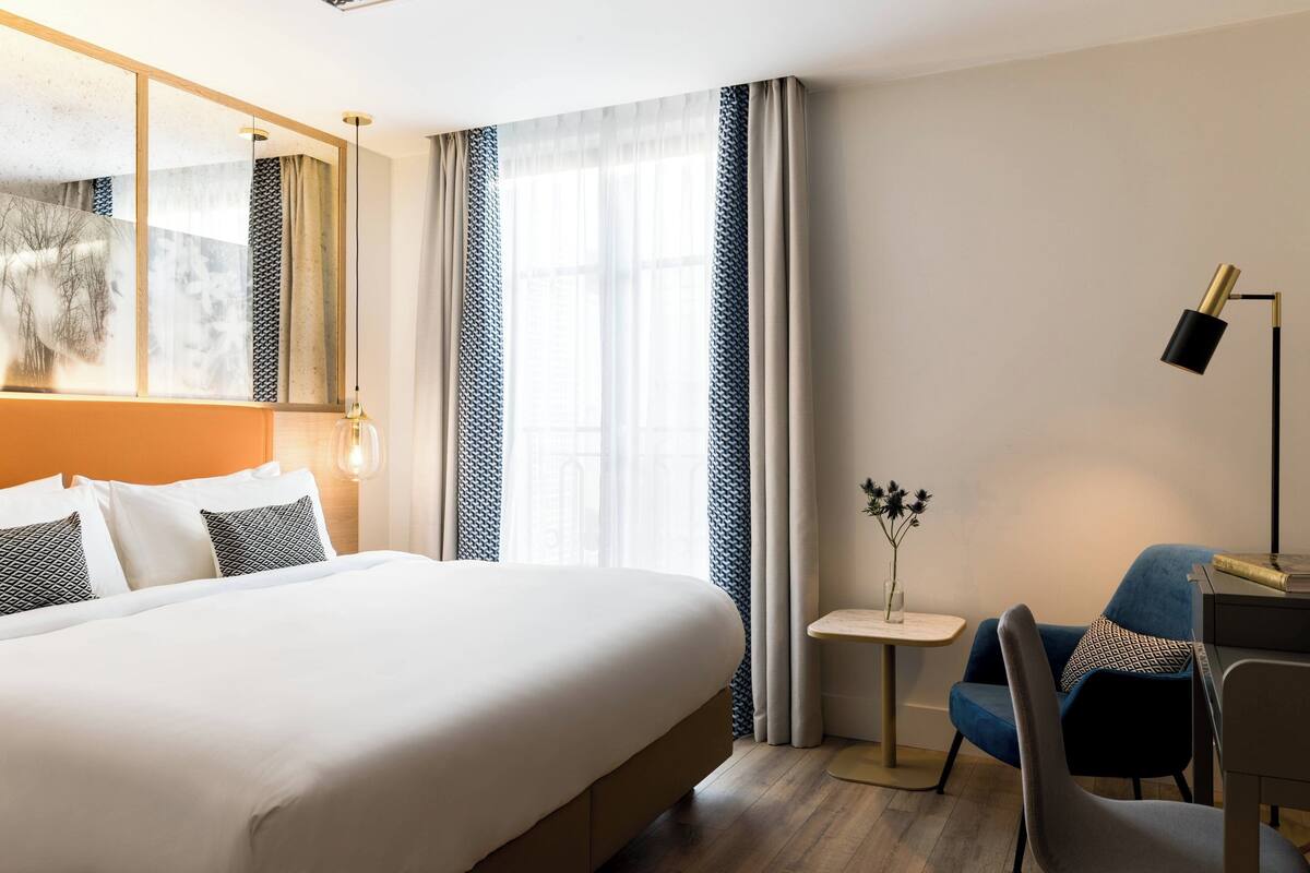 A hotel room with a large bed, a desk, a chair, a small table with a vase, and a lamp.