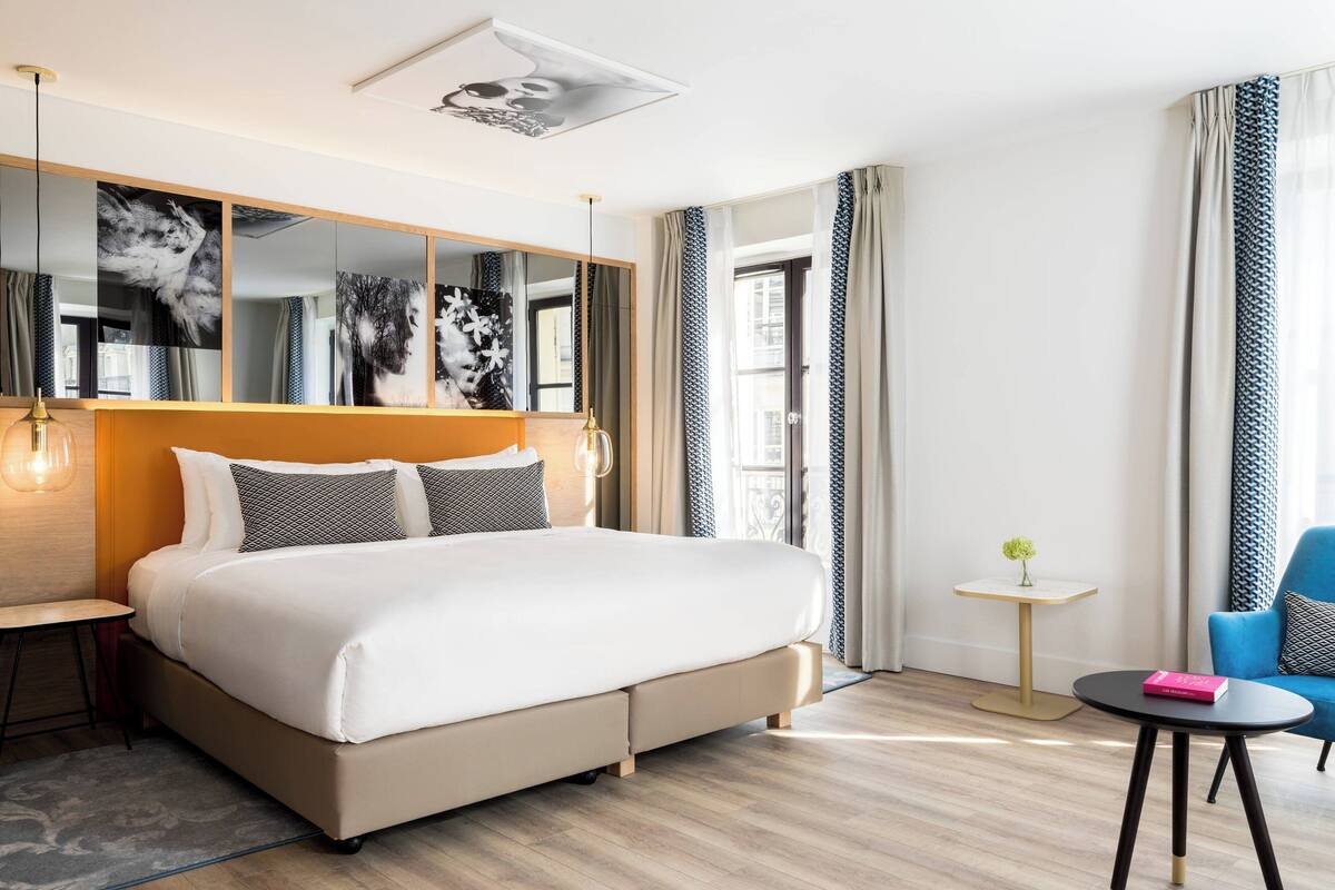 A modern hotel room with a large bed, a small table, a chair, and a mirror with artwork.